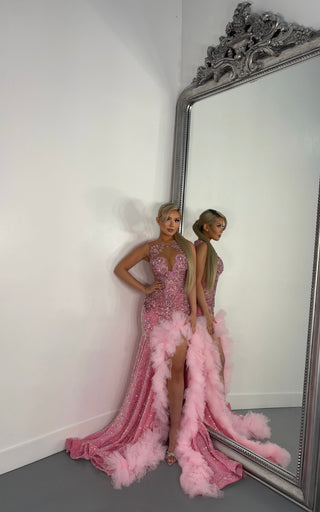 A glamorous woman in a sparkling pink gown from Alglist with feathered accents stands elegantly beside an ornate mirror against a white background