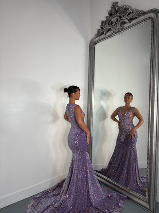 A woman in a shimmering purple evening gown poses elegantly in front of a large ornate mirror, showcasing the dress's intricate details