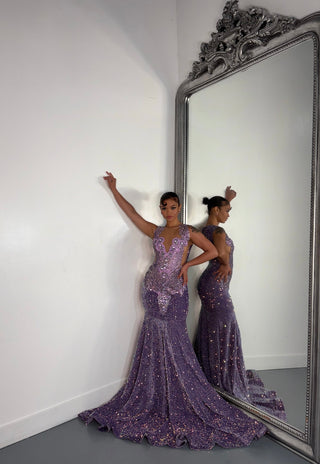A model poses in a shimmering purple gown with intricate details, reflected in an ornate mirror against a plain white wall