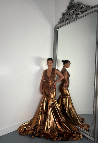 A person models a stunning, form-fitting bronze evening gown with intricate beadwork, standing beside a decorative mirror
